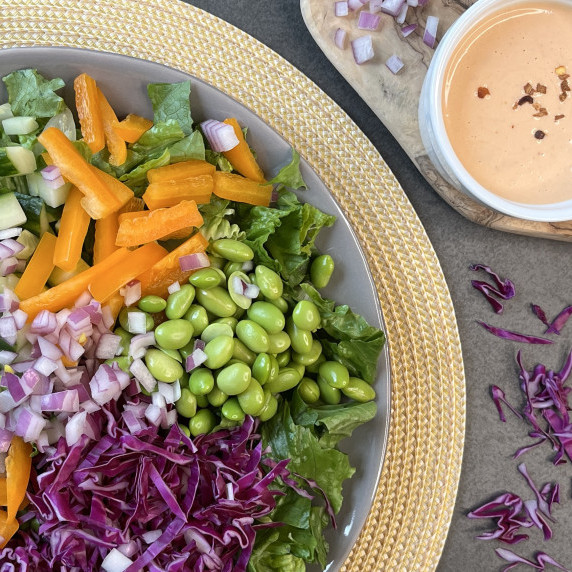 Lettuce, red cabbage, bell pepper, onion and edamame on a gray plate with dressing in a white bowl.