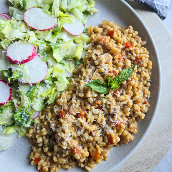 Barley risotto next to a fresh green salad with radishes on a white plate.