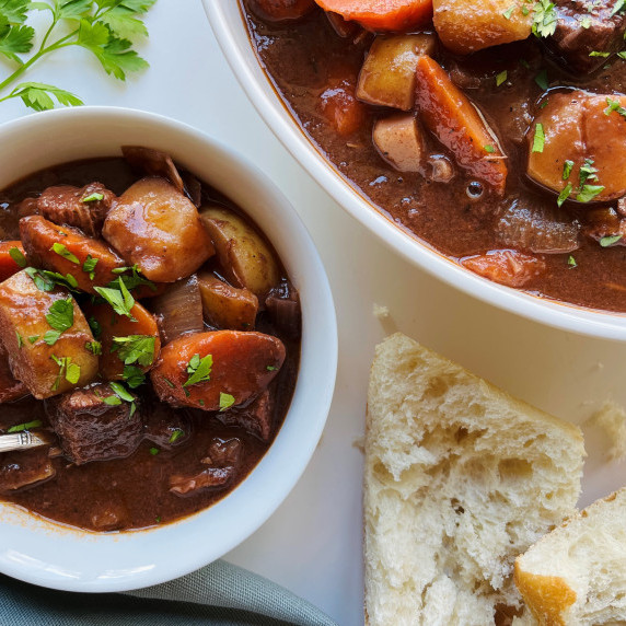 Beef stew in a white bowl with torn crusty bread on a white counter.