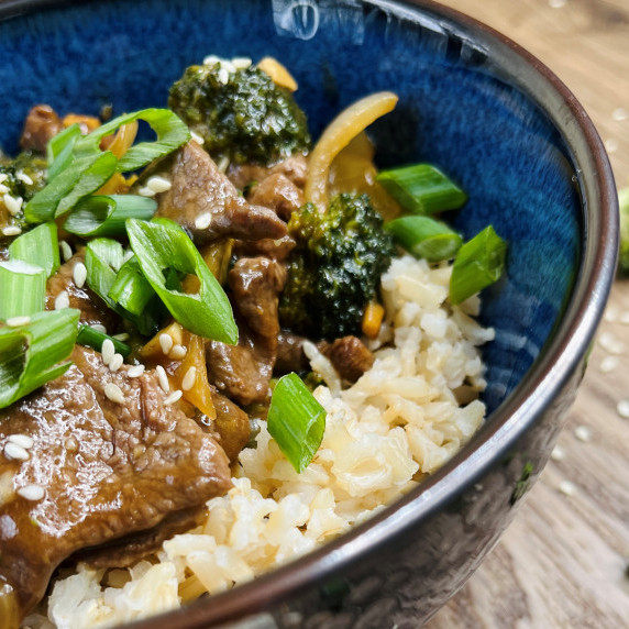 Beef and broccoli stir fry over brown rice in a blue bowl on a wood cutting board.