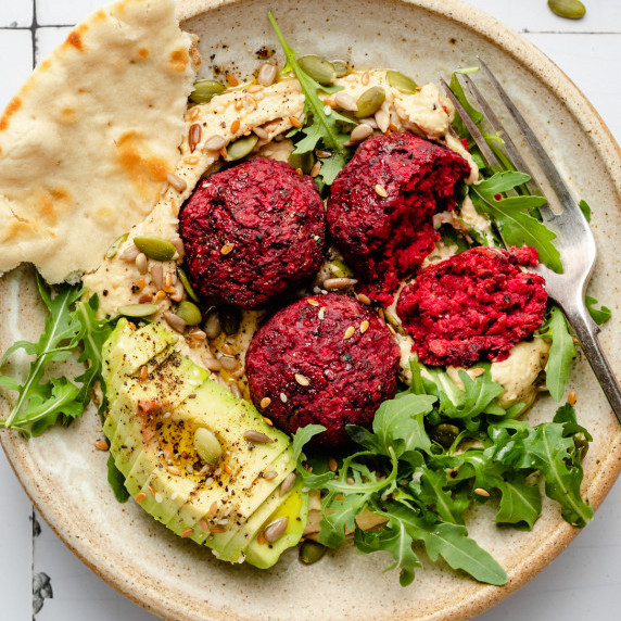 A bowl of beetroot falafel on a bed of hummus with avocado, arugula and a torn flatbread with a fork