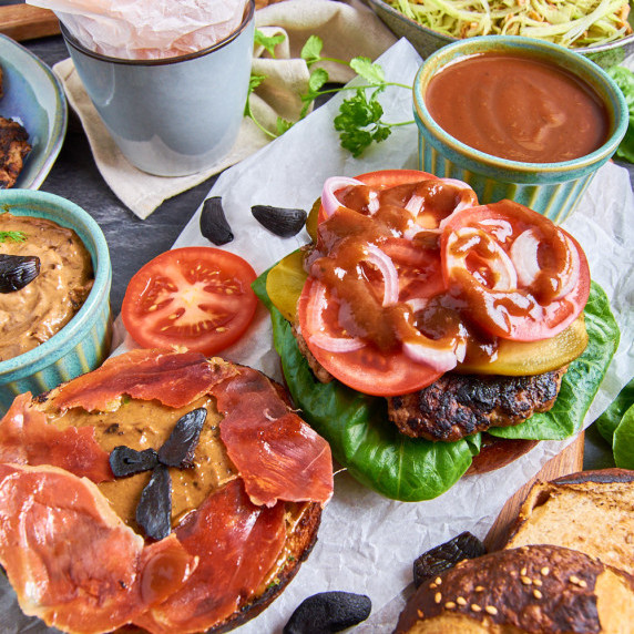 An open Pretzel Burger with Black Garlic Aioli surrounded by various toppings and sides.