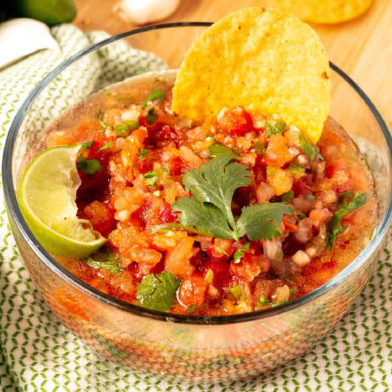 Tomato salsa in a bowl from the side with a chip and cilantro on top, a side of lime on the edge.
