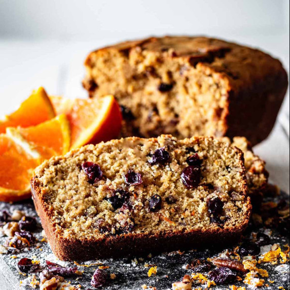 Close up of a slice of cranberry orange bread with sliced oranges and loaf in the background.