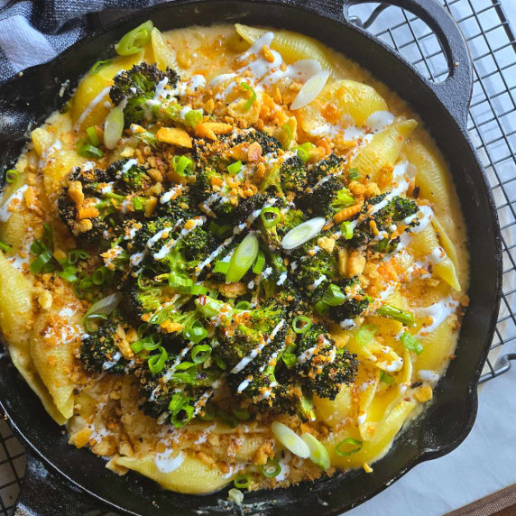 A black skillet full of golden, cheesy pasta topped with green broccoli, white sour cream, and onion