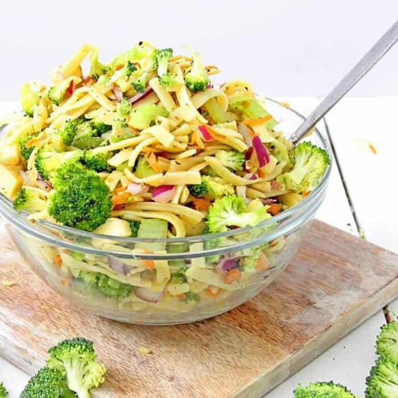 Glass bowl of broccoli apple pasta salad on a wooden cutting board.