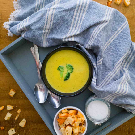 Broccoli soup in a black bowl on a grey tray with croutons, two spoons, a towel and a little jar.