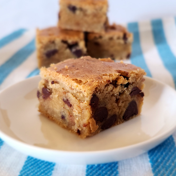 Brown butter blondie on a white plate in the foreground, more blondies on a striped cloth behind.