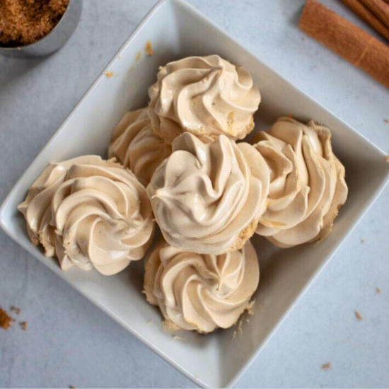 Light brown sugar meringues piped into swirls and piled in a square white bowl with cinnamon sticks.