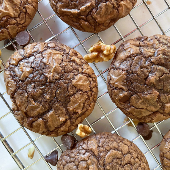 Brownie cookies on a wire cooling rack with chocolate chips and walnuts on a white counter.