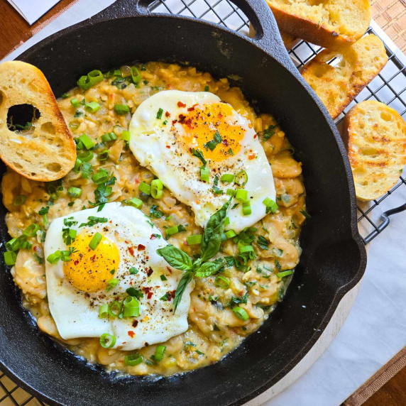 A skillet full of creamy beans, gorgeous fried eggs, green herbs, and golden grilled baguette.