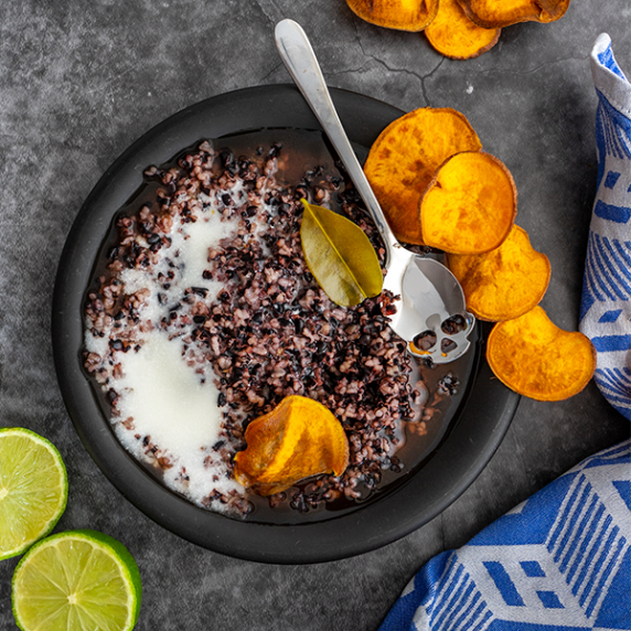 A bowl of black rice pudding with coconut milk and sweet potato chips and a skull shaped teaspoon.