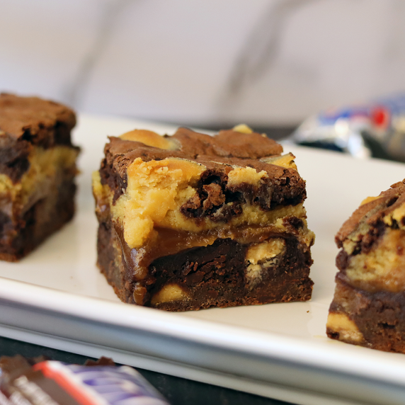 Candy Bar Cheesecake Brownies arranged on a narrow, rectangular, white platter