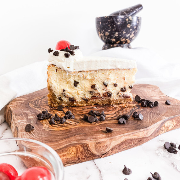 Slice of cannoli cheesecake on an olive wood board with chocolate chips on a white counter