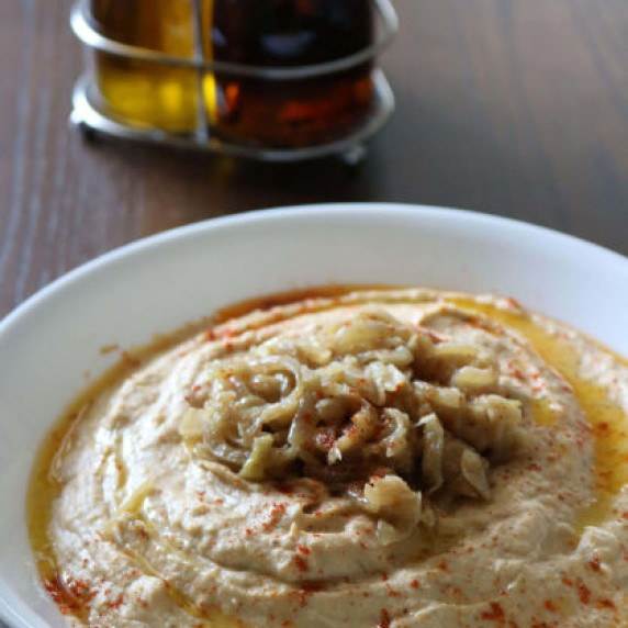 A white bowl of hummus topped with olive oil and caramelized onions on a dark table.