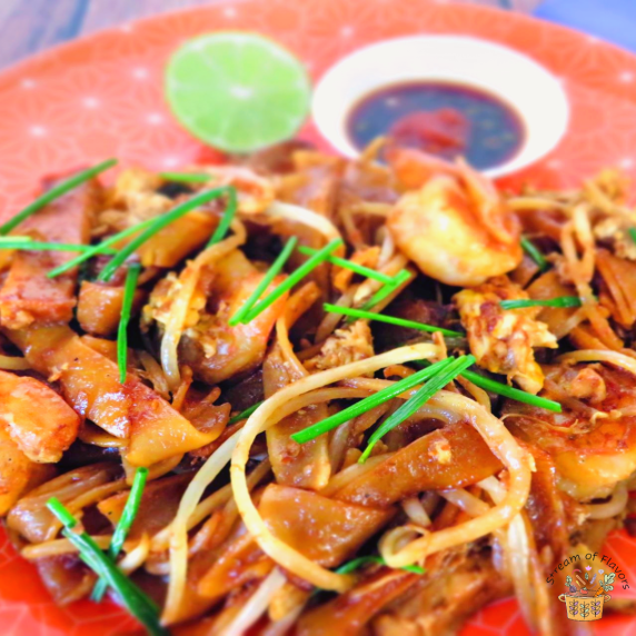Char Kway Teow with shrimp, beansprouts, eggs, and tofu on an orange plate with lime and sauce