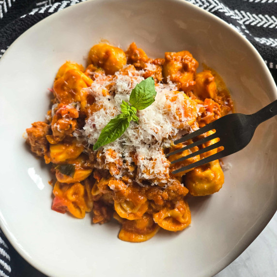 A white bowl full of orange hued tortellini with white cheese and green basil.