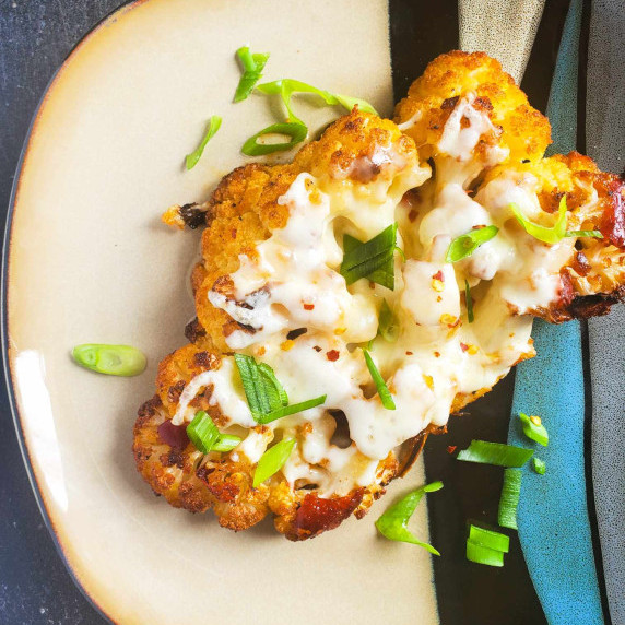 A blanket of white cheese on top of a roasted cauliflower against a multicoloured plate.