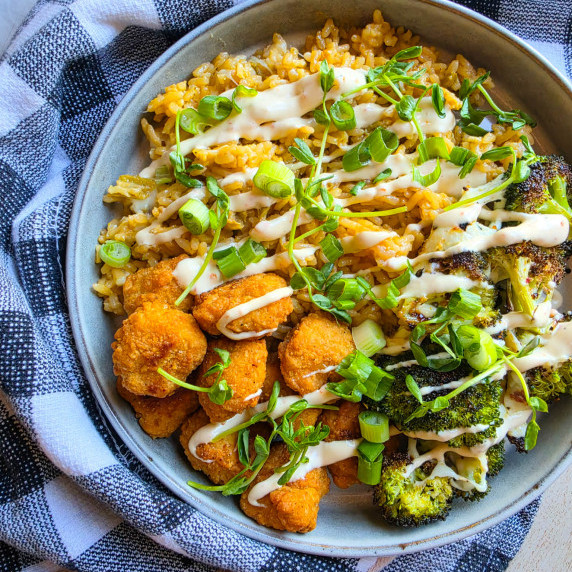 Golden chik'n, green broccoli, white sauce drizzle, and greens against a checkered towel.