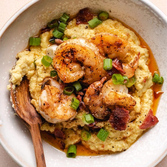 Seared shrimp on a bed of cheesy grits with sauce and spring onions in white bowl with wooden spoon