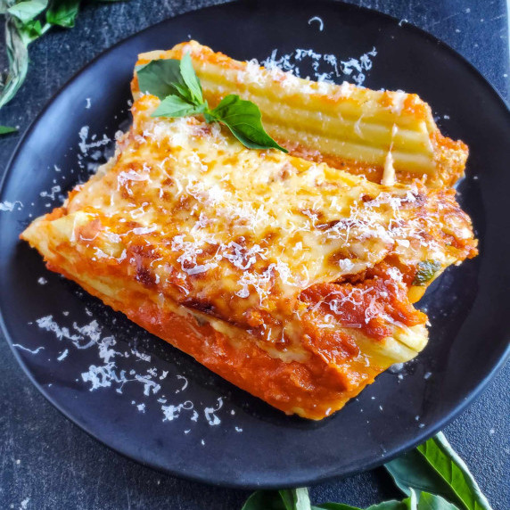 Cheesy manicotti dressed in red sauce on a black plate against a black background.
