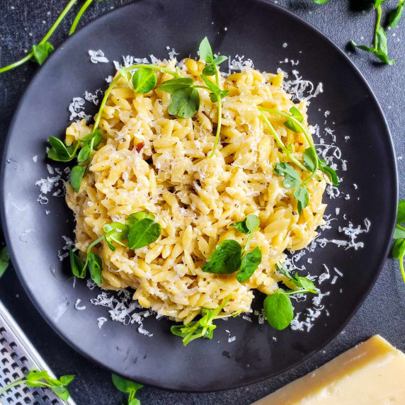 A cheesy pile of orzo, garnished with green pea shoots on a black plate.