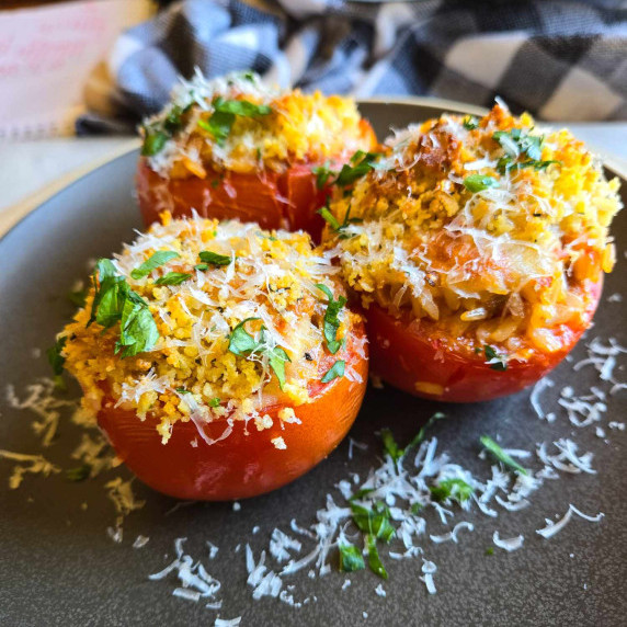 3 stuffed vine tomatoes with a golden brown crumb topping and some green herbs on a matte plate.
