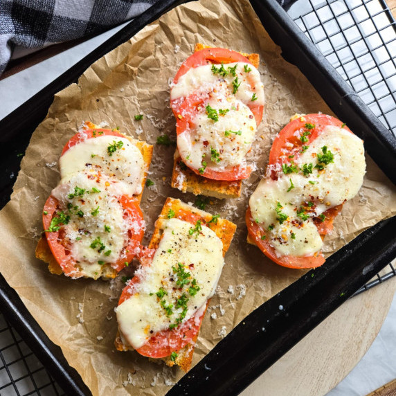 A parchment lined baking sheet with cheesy, golden baguettes with red tomatoes and pops of green