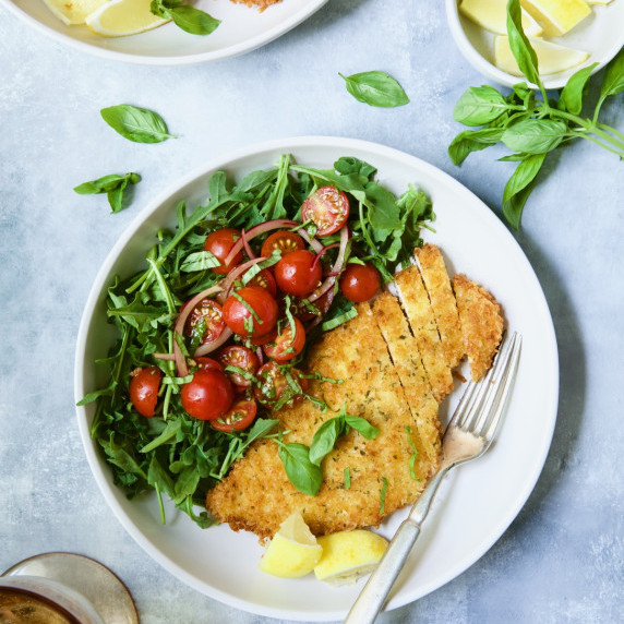 Chicken Milanese with cherry tomato and arugula salad on white plate on blue background.
