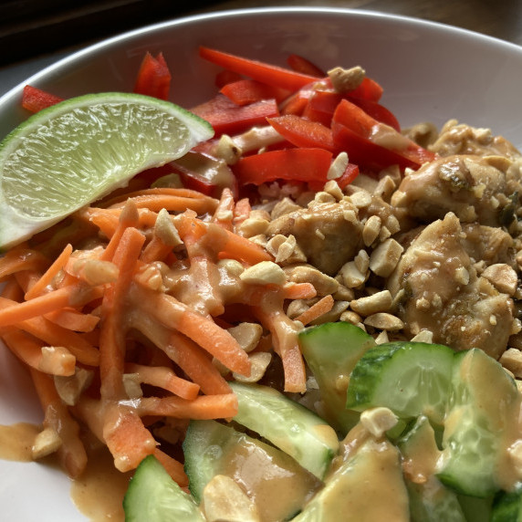 Chicken, cucumbers, shredded carrot, red pepper in a white bowl with peanut dressing.