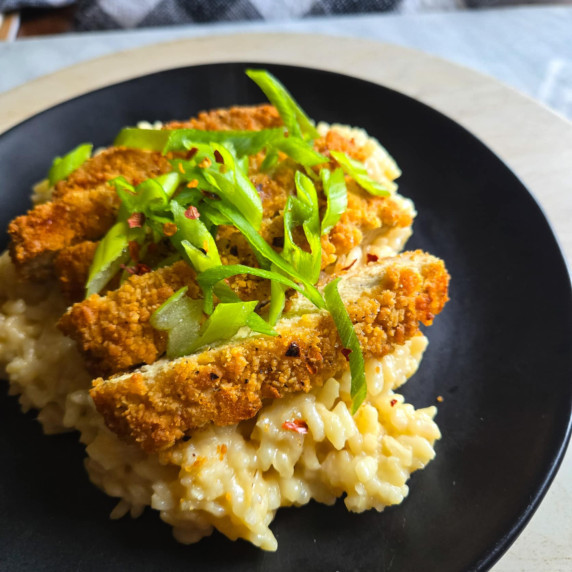 Golden brown chik'n nestled in to creamy risotto with green onions on a black plate.