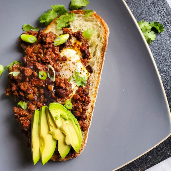Grilled toast smothered in chili and avocado against a grey plate.