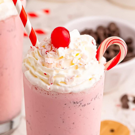 A pink milkshake topped with whipped cream, a red cherry, a candy cane, and a red-striped straw.