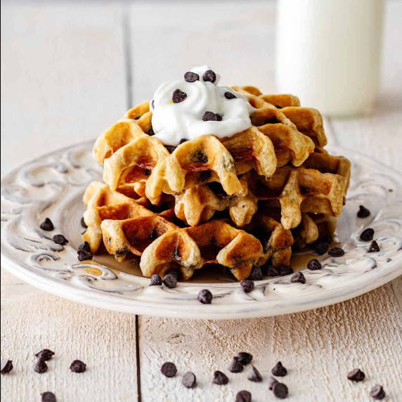 Stack of chocolate chip waffles topped with chocolate chips, whipped cream, and maple syrup.