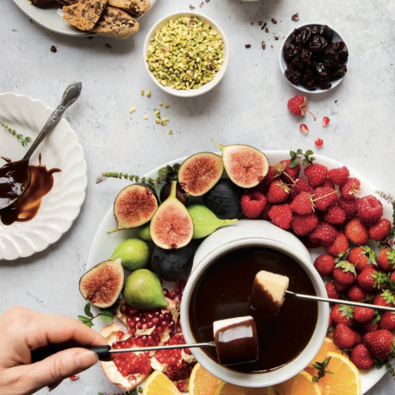 Chocolate fondue with fruit, cookies, and nuts; a hand dips a marshmallow.