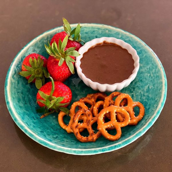 Chocolate pumpkin seed butter in white bowl on green plate with whole strawberries and pretzel twist