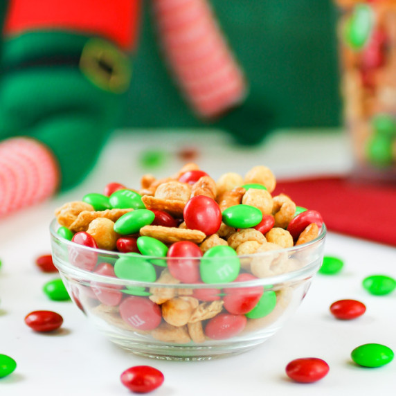 Side view of Elf Nibbles in a clear bowl with M&M's sprinkled around with Christmas decorations.