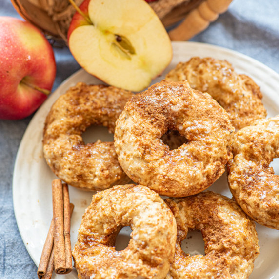 Apple cinnamon donuts stacked on a plate with apples and cinnamon sticks nearby