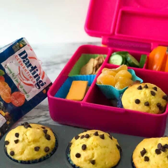 A child's lunchbox full of food with muffins in a pan in front and a bag of clementines in the back.