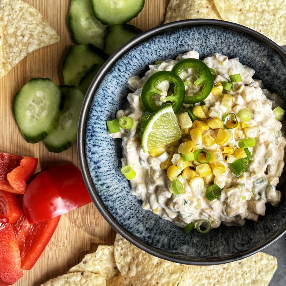 Blue bowl filled with corn dip on a wooden board with corn chips and cucumber and red pepper slices.