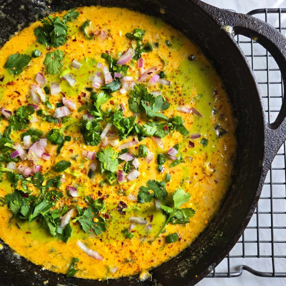 A bright yellow, vibrant corn queso skillet garnished with greens and red onions.