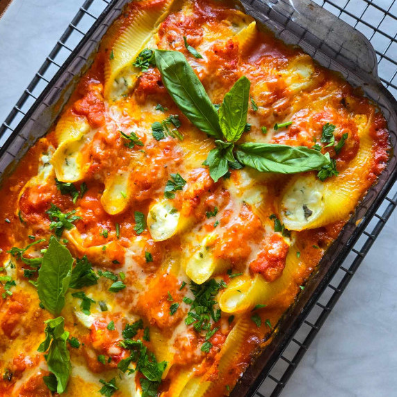 A dish full of stuffed shells smothered in a vibrant red pepper sauce and melty cheese with herbs.