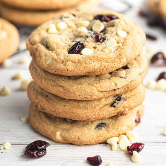 Four cranberry white chocolate cookies stacked with extra chocolate and cranberries scattered nearby