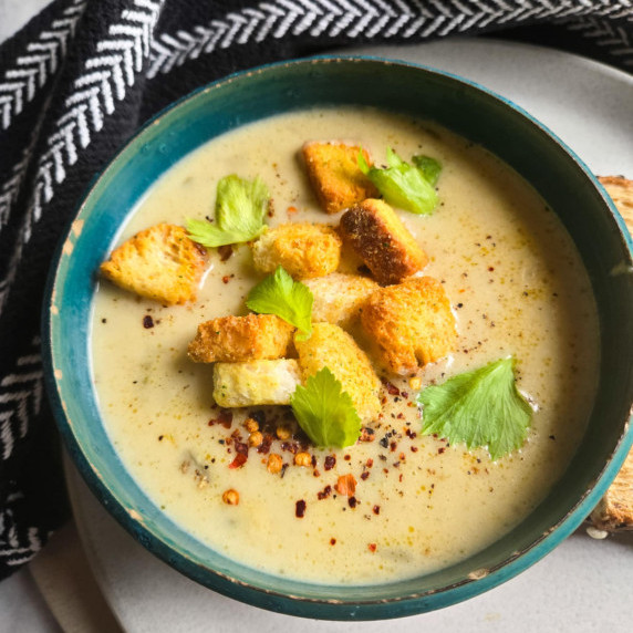 A blue bowl full of creamy soup with golden croutons and green celery leaves.