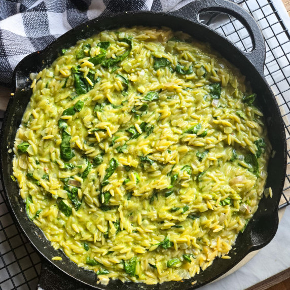 A black skillet full of green pesto orzo with a black and white checkered towel in the background