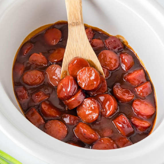 Sliced BBQ smoked sausage bites in a BBQ sauce in a white crockpot being scooped with a wooden spoon