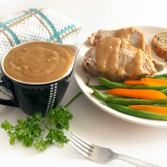 Brown gravy in a small gravy boat with a plate of pork, gravy and vegetables next to it.