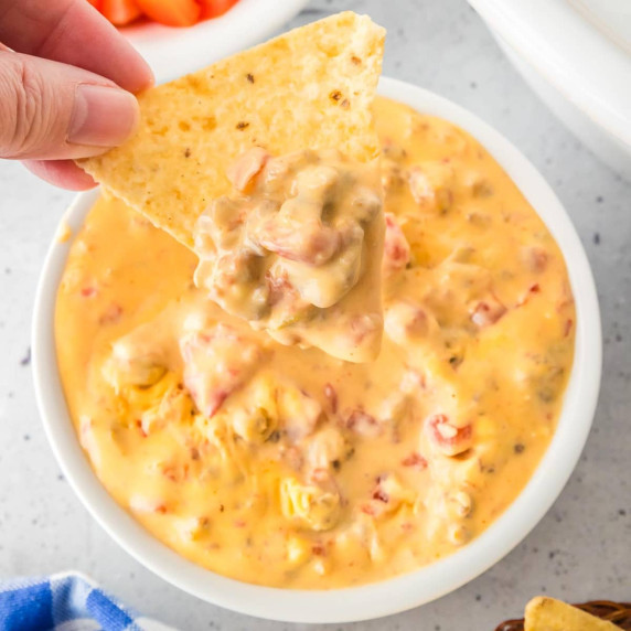 A hand holds a tortilla chip dipped in a bowl of creamy Rotel dip next to a slow cooker on a counter