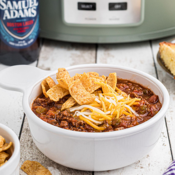 A bowl full of chili in front of a crock pot with a bottle of beer next to it.