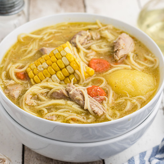 A close up shot of a bowl of cuban chicken soup with a corn cob, potato and some carrots visible.